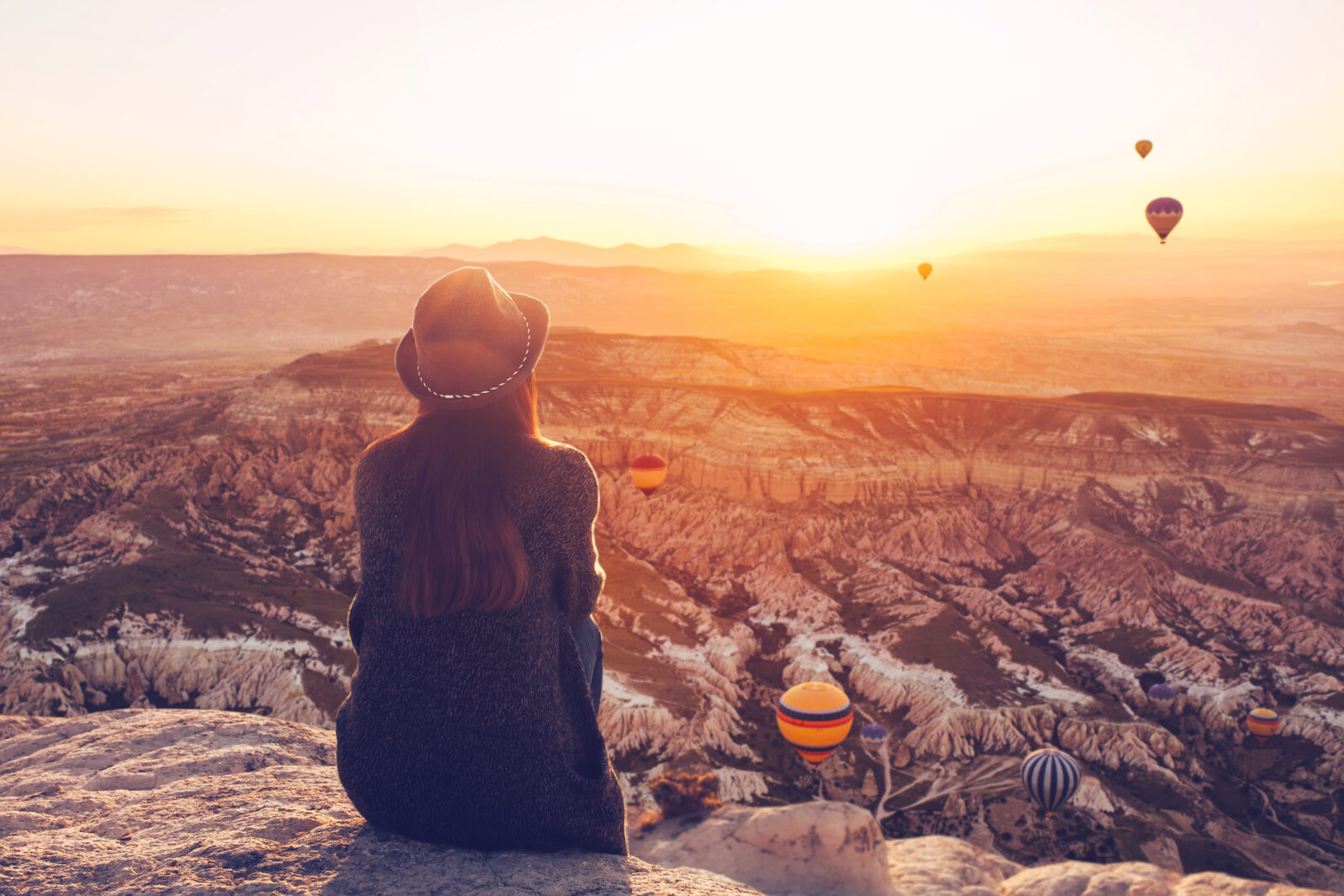 A girl in a hat on top of a hill in silence and loneliness admires the calm natural landscape and balloons.