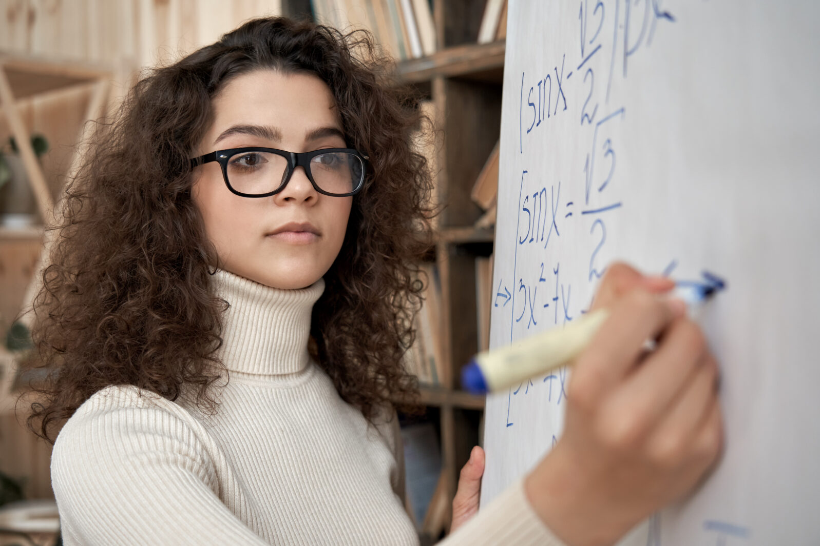Young serious female latin math school teacher wearing glasses holding writing equation on whiteboard in classroom. Hispanic university college tutor, graduate student learning, teaching during class.