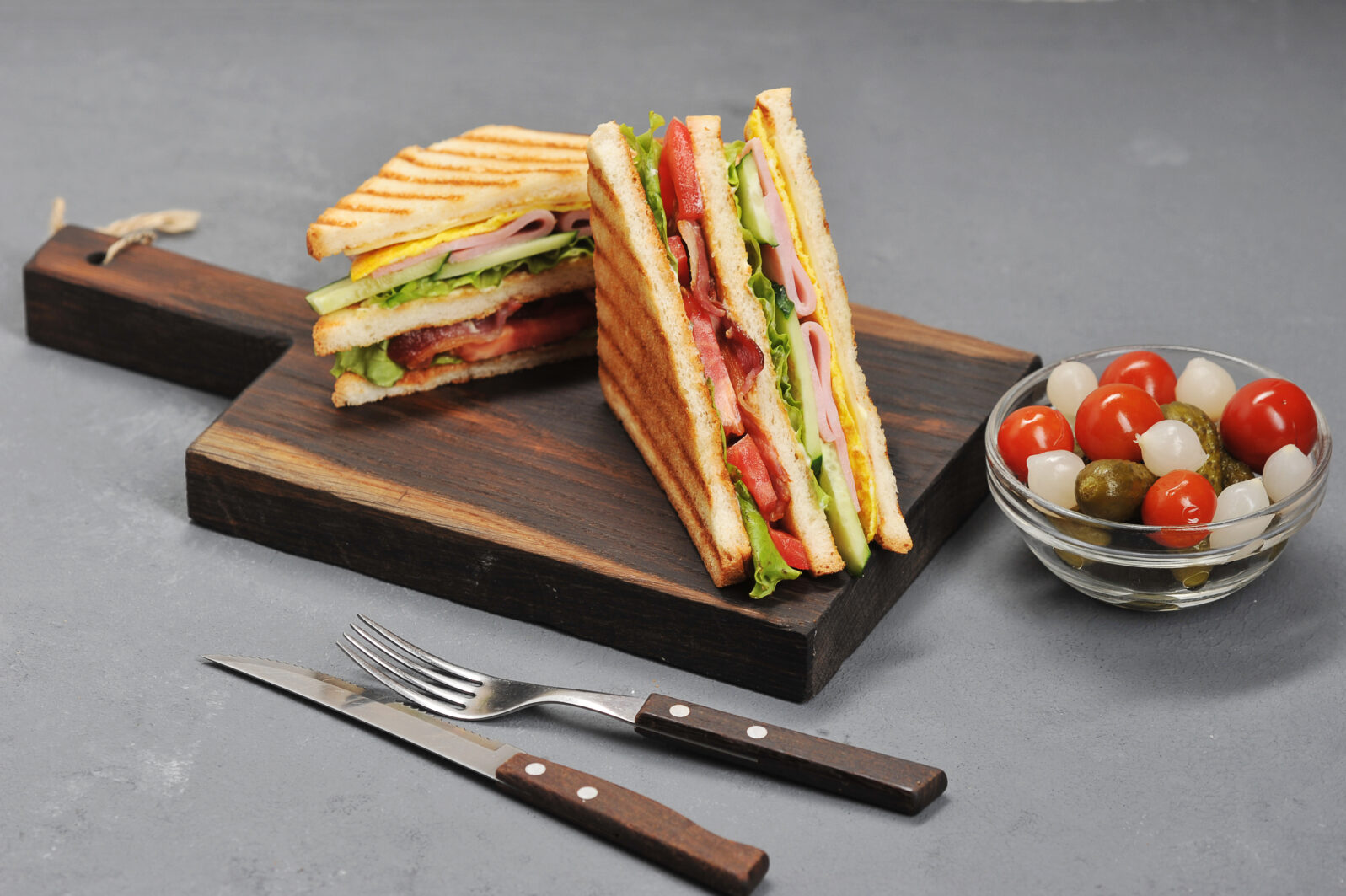 Classic club sandwich with ham and bacon on a wooden board. Next cup is a mixture of pickled miniature onions, gherkins, tomatoes. Gray background. Close-up.