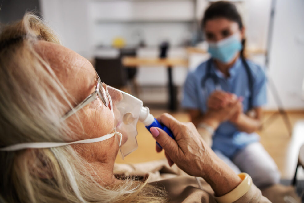 Closeup of ill blond senior woman sitting at home and taking oxygen from respirator while nurse with protective mask on sitting next to her and holding her hand during corona virus.