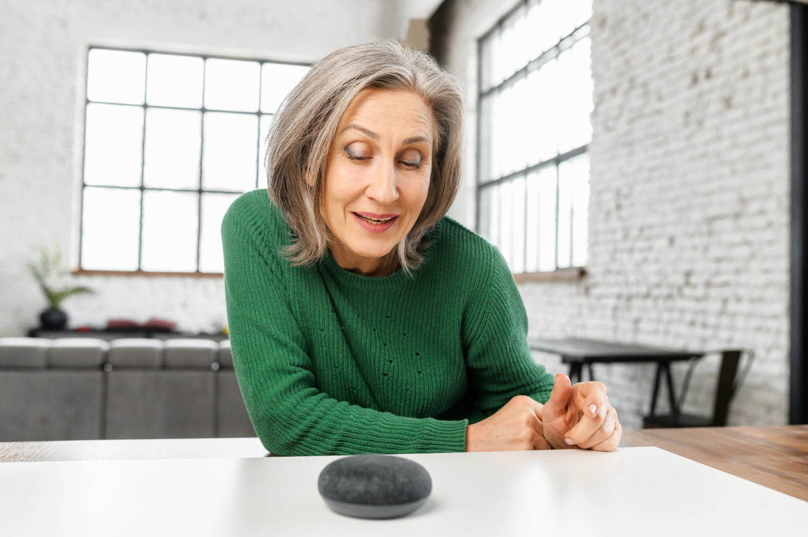 woman with gray hair in green jumper talking to the digital virtual assistant at home, asking a question or requesting to switch music. Smart AI speaker concept and voice command control