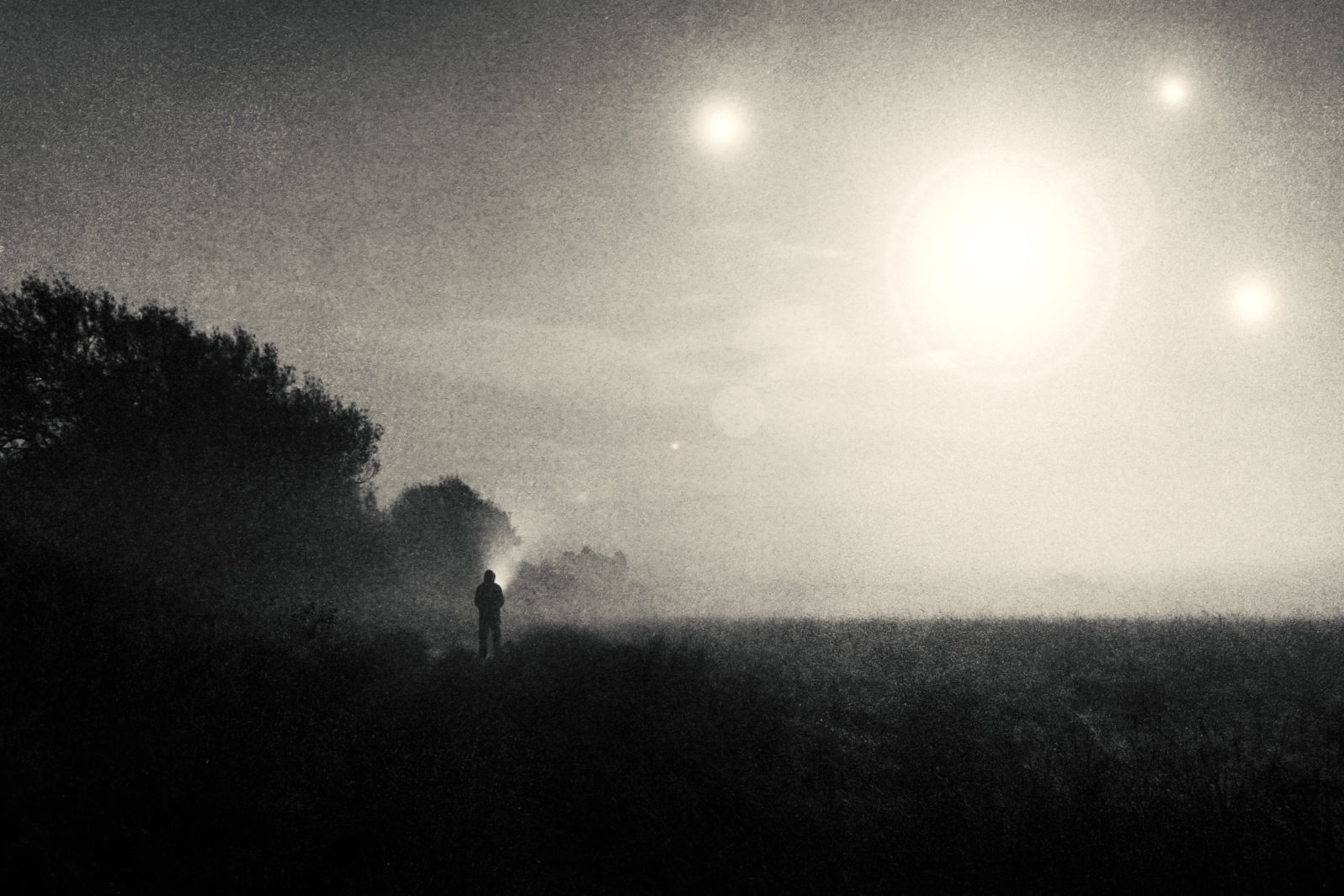 A moody science fiction concept, of a figure standing in a field with UFO lights glowing in the sky. On a foggy spooky night. With a vintage, grunge edit