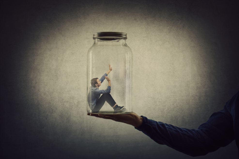Business concept with a scared tiny man trapped inside a glass jar held by his gigantic boss hand. Surreal nightmare, helpless captive employee victim of abuse at work. Workplace bullying and conflict
