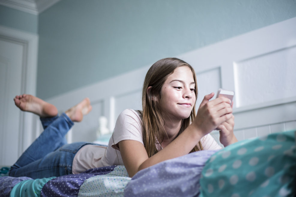 Pre-adolescent teen girl texting on a smartphone lying in bed at home. Candid indoor photo withFocus on the foreground and copy space