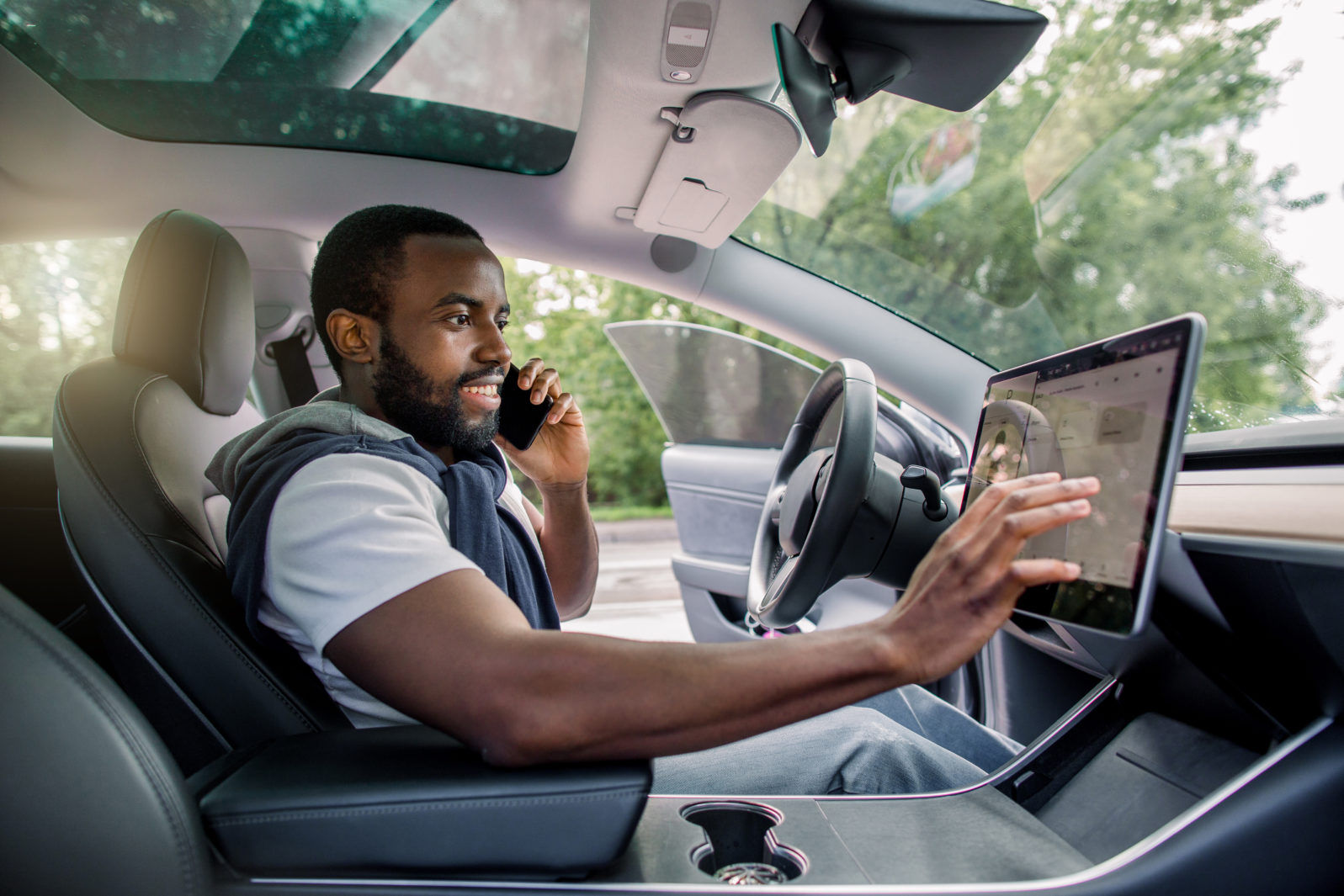 Handsome bearded African man touches the touchscreen in his new high-tech electric vehicle while while talking by phone and smiling. Self driving vehicle concept
