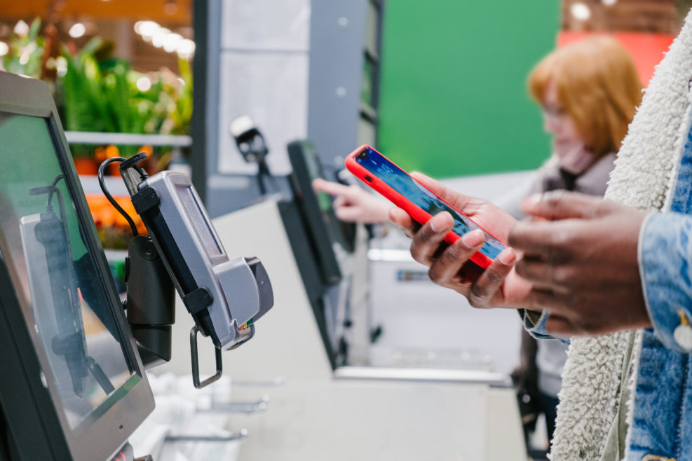 Black male person in warm denim jacket uses smartphone to pay for purchase at self-checkout point in supermarket close view
