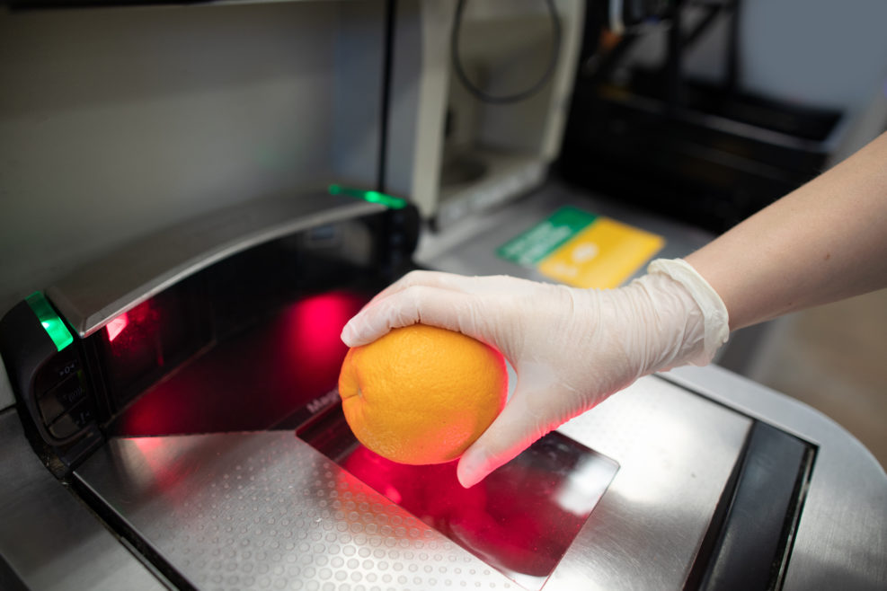 A hand in disposable latex glove scanning an orange at a grocery store self-checkout station. People wearing gloves to protect themselves from covid-19 pandemic.
