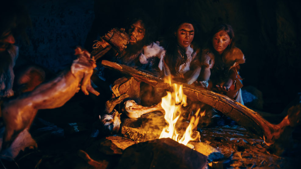Neanderthal or Homo Sapiens Family Cooking Animal Meat over Bonfire and then Eating it. Tribe of Prehistoric Hunter-Gatherers Wearing Animal Skins Eating in a Dark Scary Cave at Night