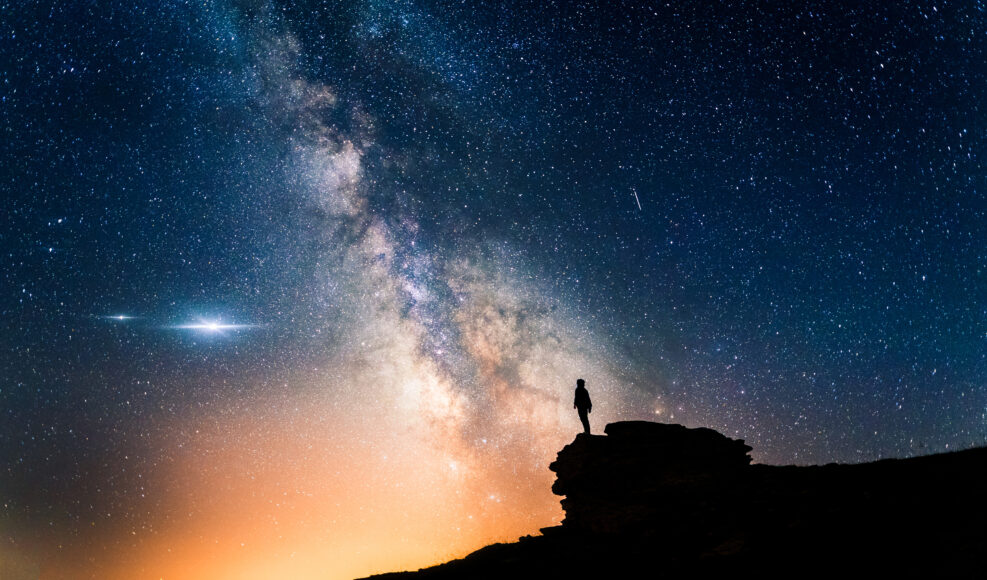 Man standing in a high place looking up in wonder to the Milky Way galaxy. Small silhouette of a man under the Milky Way and the magical starry sky. Concept of human smallness.