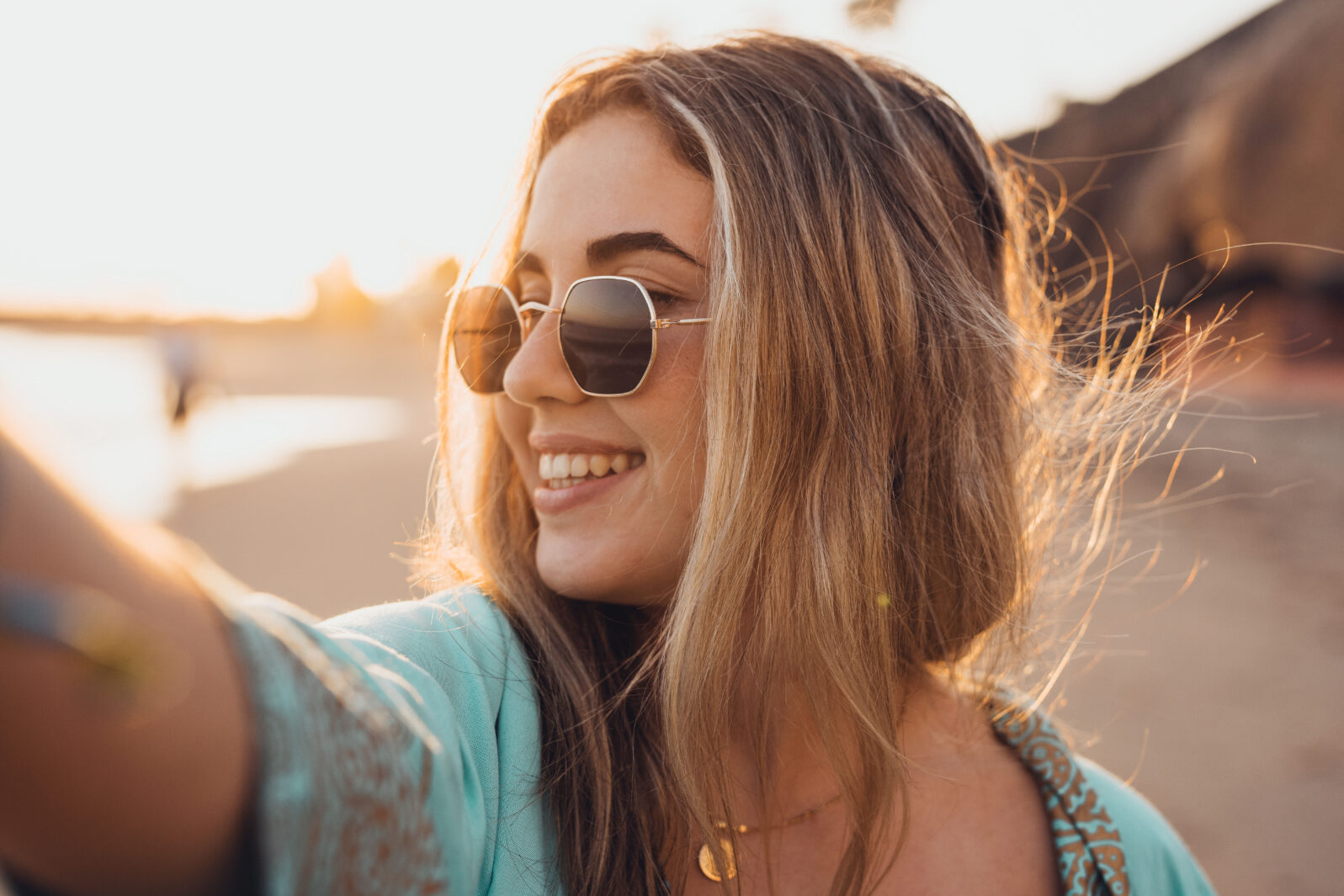 Portrait close up of one young beautiful attractive blonde young girl holding camera and taking selfie picture at the beach enjoying outdoors free time with sunset in the background.