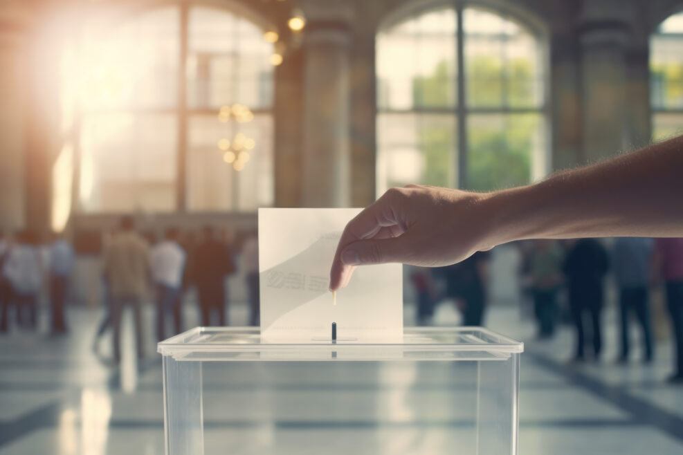 a hand drops a ballot into a transparent plastic ballot box on election day at a polling station. the will of the voters. free elections. Generative AI