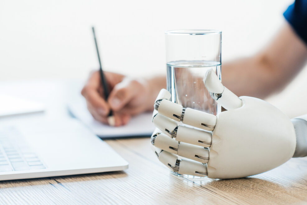 selective focus of robotic arm holding glass of water and person taking notes at wooden table