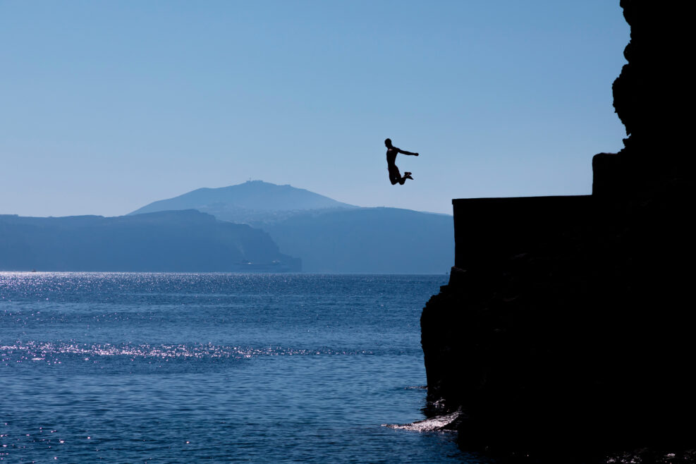 Tourists jumping off a large rock ledge in Amoudi Bay on Santorini Island in Greece.