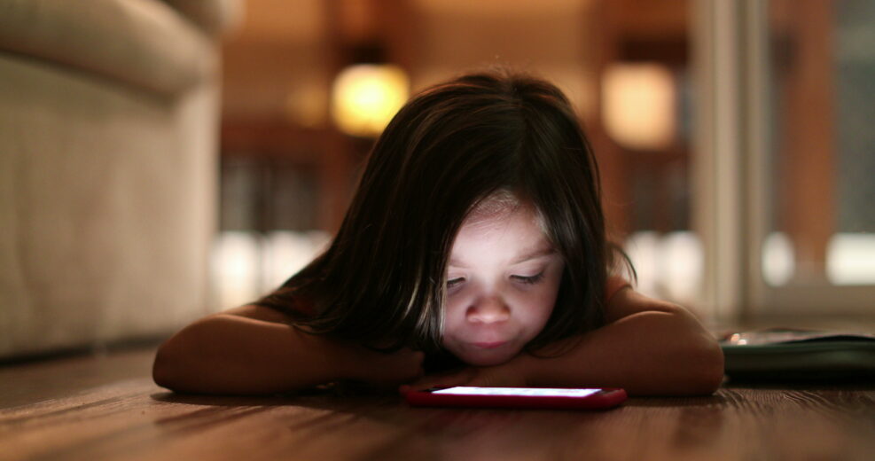 Child watching cellphone screen at night. Blue light from smartphone device glowing on little girl face
