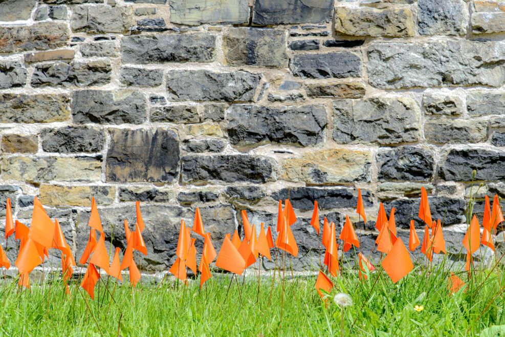 Small orange flags places in grass in memory of the thousands of Indigenous children that died in Canada's residential school system. Wide view.