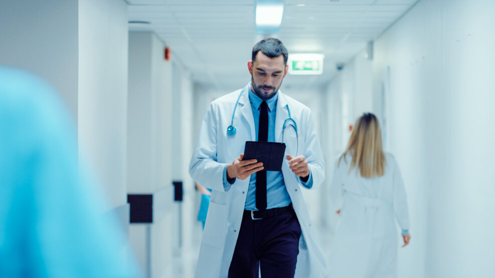 Determined Handsome Doctor Uses Digital Tablet Computer while Walking Through Hospital Hallway. Modern Bright Clinic with Professional Staff.