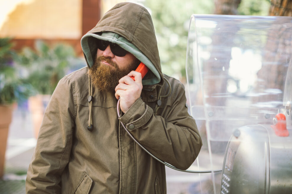 Portrait of young bearded man using public phone wearing hoodie (jacket) and black sunglasses - informant, spy, secret agent and fake news concept