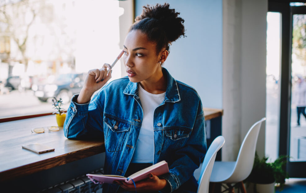 Thoughtful female blogger looking away and feeling pondering on ideas for new article at own web page, pensive woman sitting in cafeteria with textbook in hand and thinking about plan for future trip
