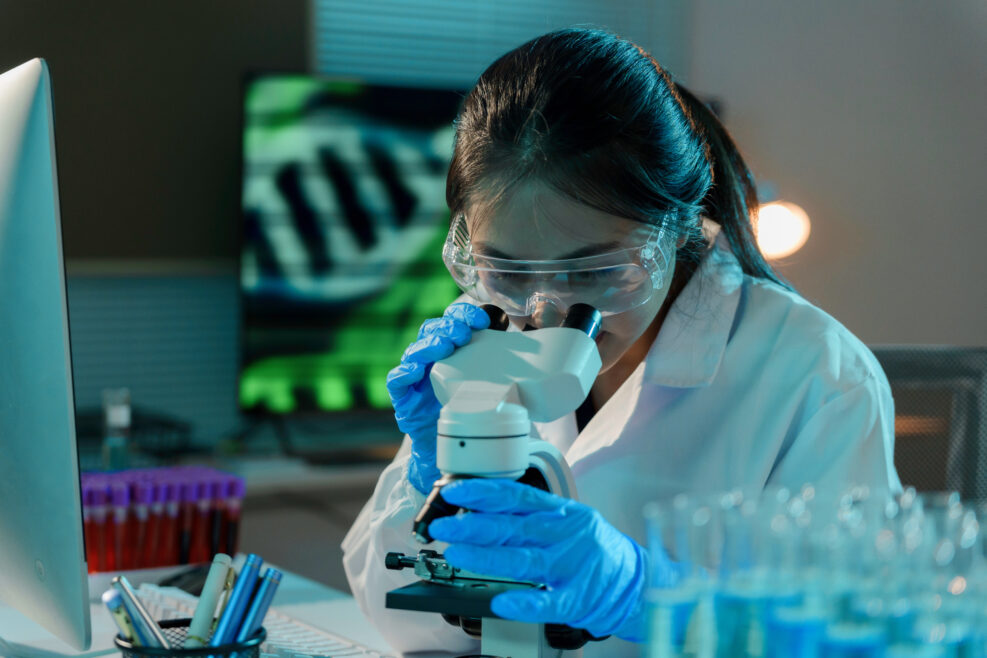 Young scientist wearing a lab coat and protective glasses is adjusting a microscope while working late in a laboratory