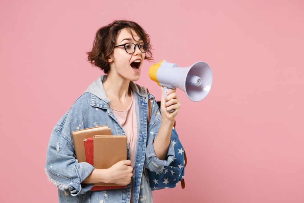 Amazed young woman student in denim clothes, eyeglasses, backpack posing isolated on pastel pink background. Education in high school university college concept. Holding books, scream in megaphone.