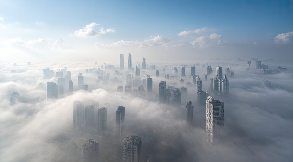 City skyline, shrouded in a thick layer of fog. Elevated aerial view reveals numerous high-rise buildings emerging from a dense cloud cover. A clear, pale blue sky is visible above the clouds