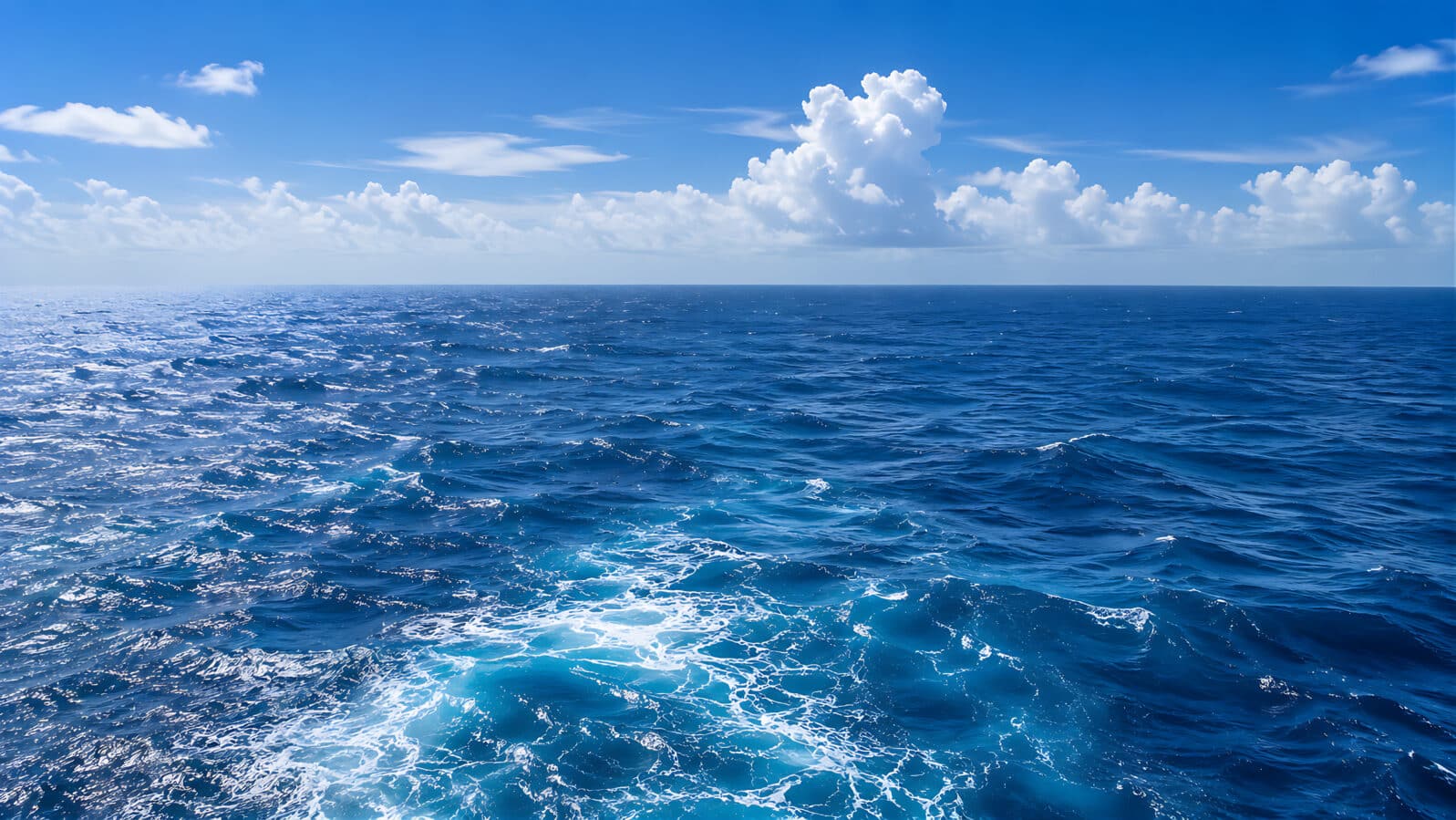 A breathtaking panoramic view of the deep blue ocean stretching to the horizon beneath a vibrant sky filled with fluffy white clouds. Gentle waves shimmer in the sunlight, creating a peaceful seascape