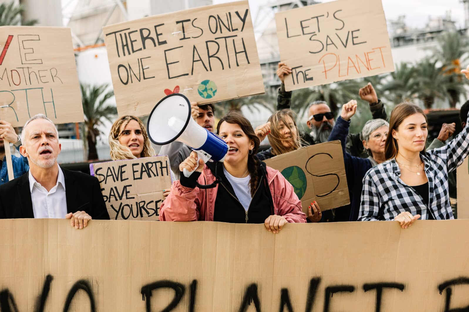 Crowd of diverse protestors demonstrating against climate change in front of factory. Global warming concept with activist holding placards and raising fists.
